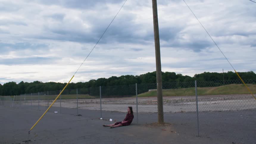 A woman sits on the asphalt against a fence