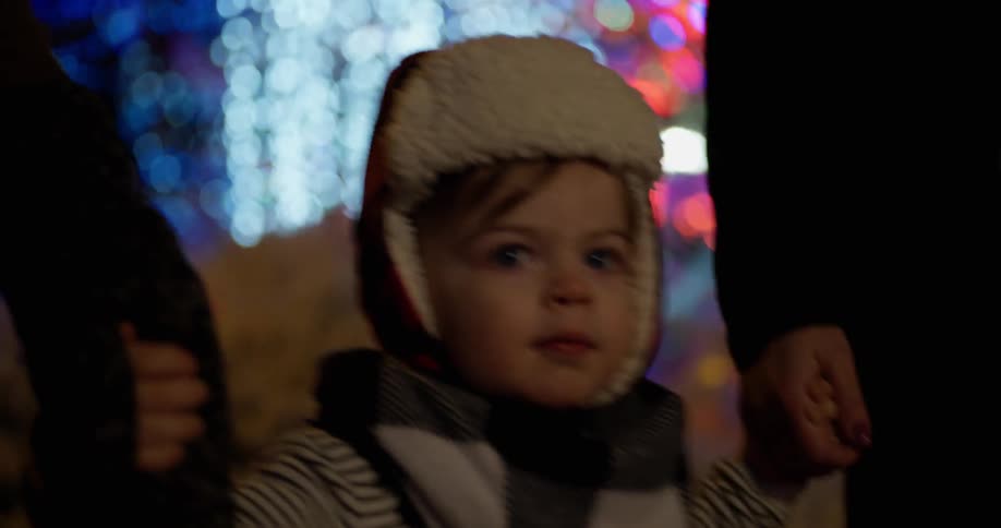 A young child is holding the hands of its parents while they all look at christmas lights