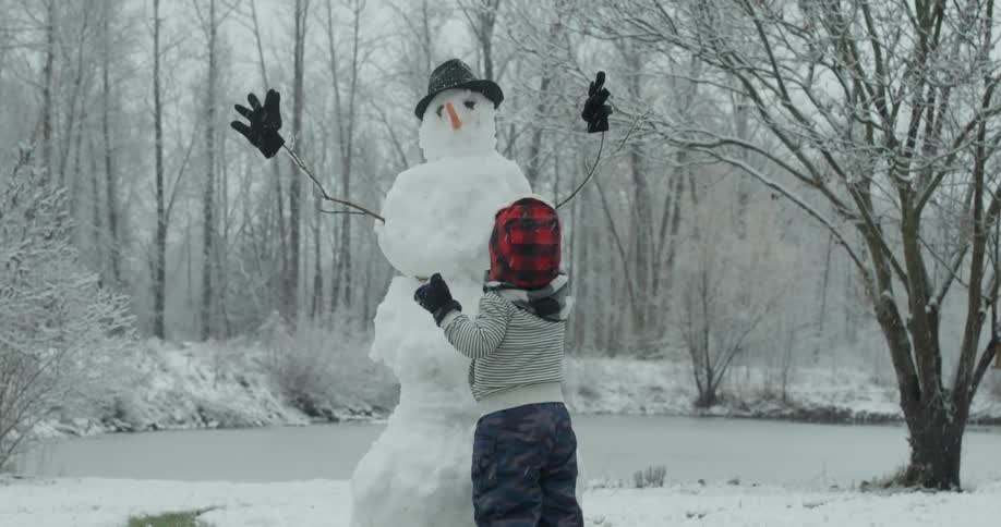 A young child stands next to a built snowman
