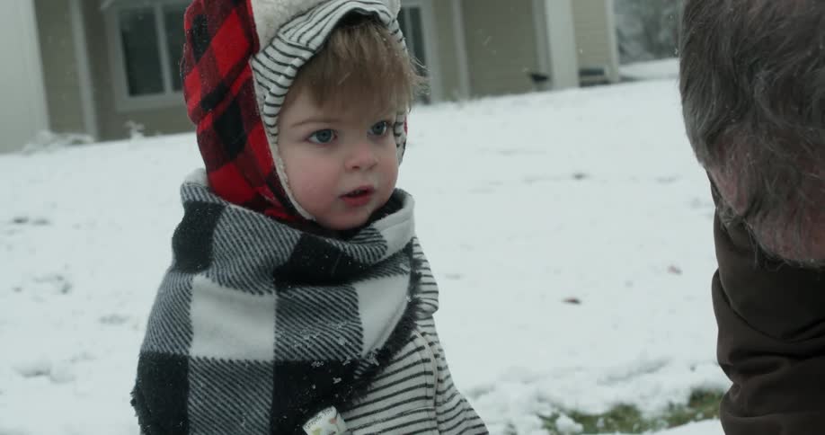 A toddler boy holding snow in his hands looks up