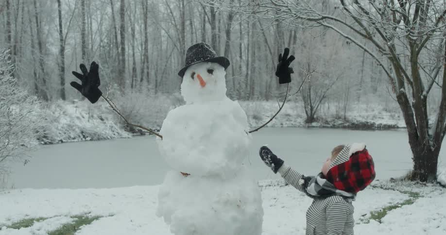 A young child stands next to a built snowman