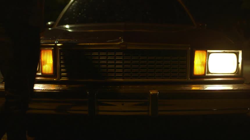 A person leans against the hood of a car at night