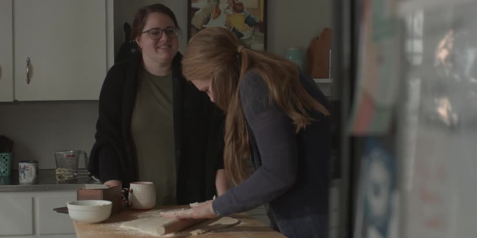 Two women talk in a kitchen while one of them rolls out dough