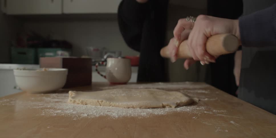 A person uses a rolling pin to flatten cookie dough as a person sprinkles flour