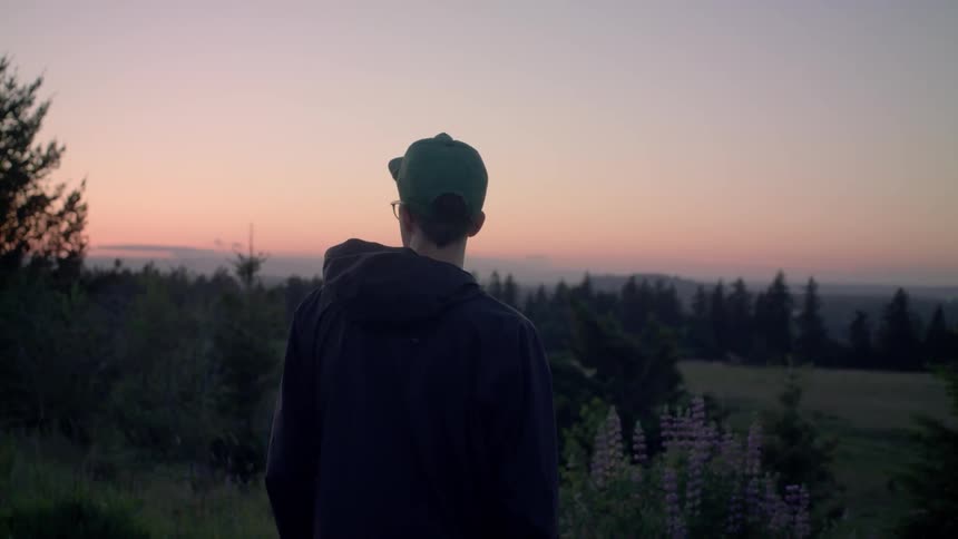 A man stands outside and looks towards tall trees