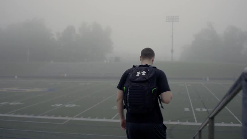 A man with a backpack is walking down the bleachers towards the football field