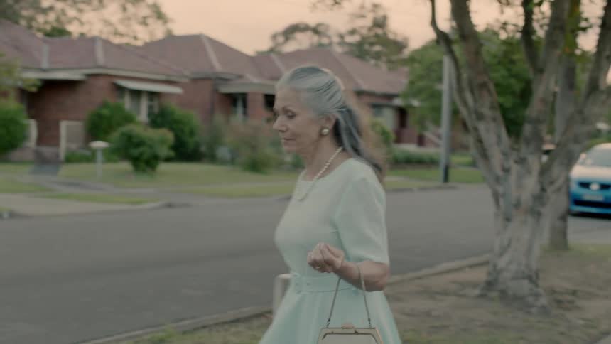 An old woman dances and twirls as she walks down a suburban sidewalk