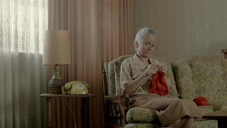An old woman sitting in a living room knitting stops and gets up