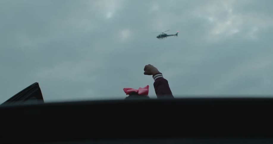 Someone wearing a pink hair bow raises their fist towards  a helicopter hovering in a cloudy sky