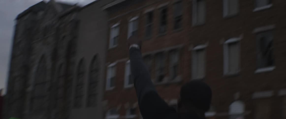 A man hanging out of a car window raises his fist up into the air as the car proceeds down the street
