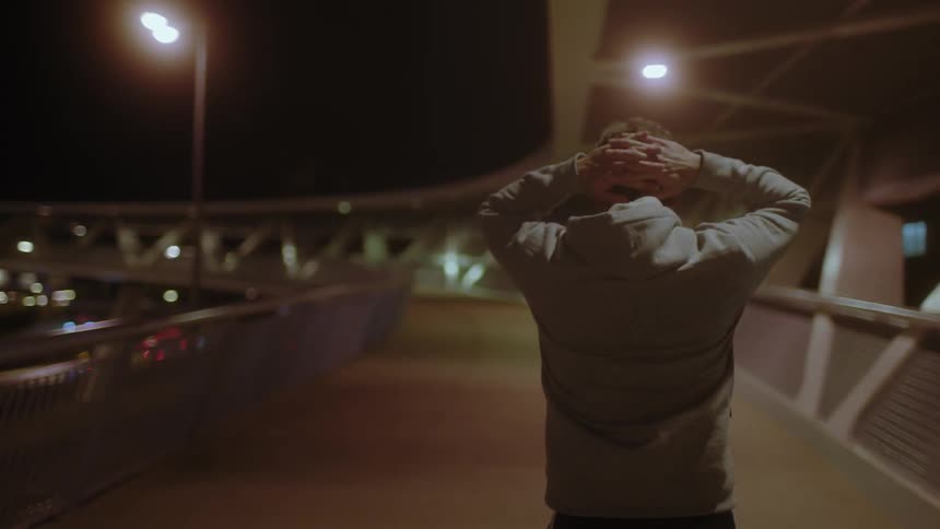Man walks on a bridge outside at night with hands on his head