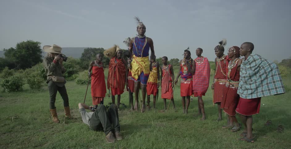 Maasai wearing costumes stand outside while one man jumps up and down and another man takes his picture