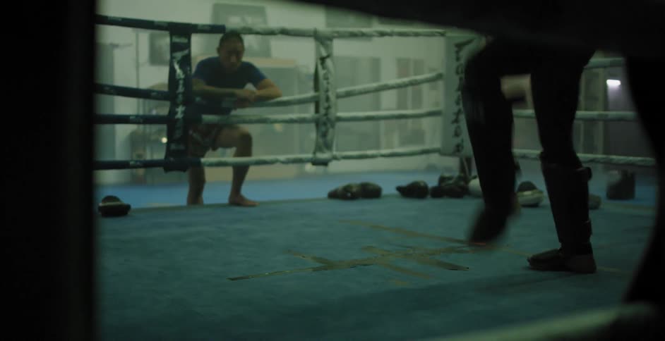 A man leans on the ropes while a kick boxer practices in a ring