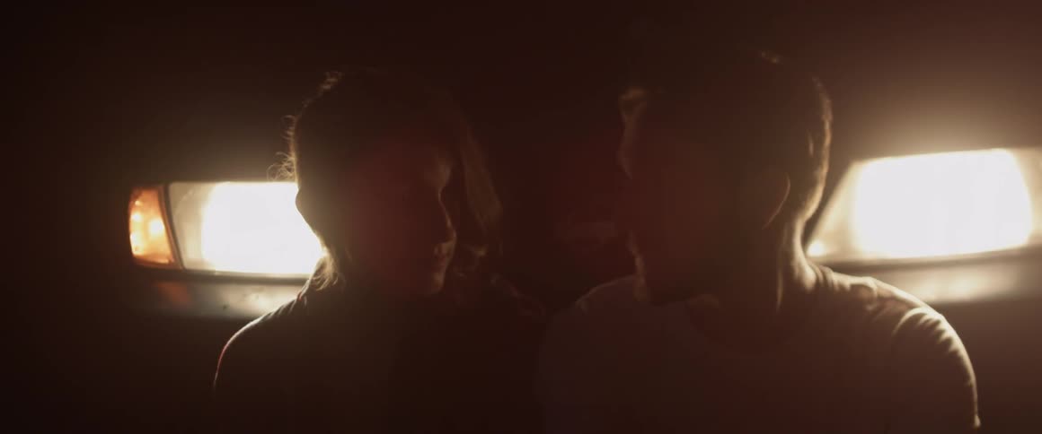 A man smokes while he and a woman sit in front of a car