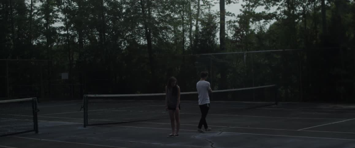 A man and woman are standing in the middle of a tennis court at dusk