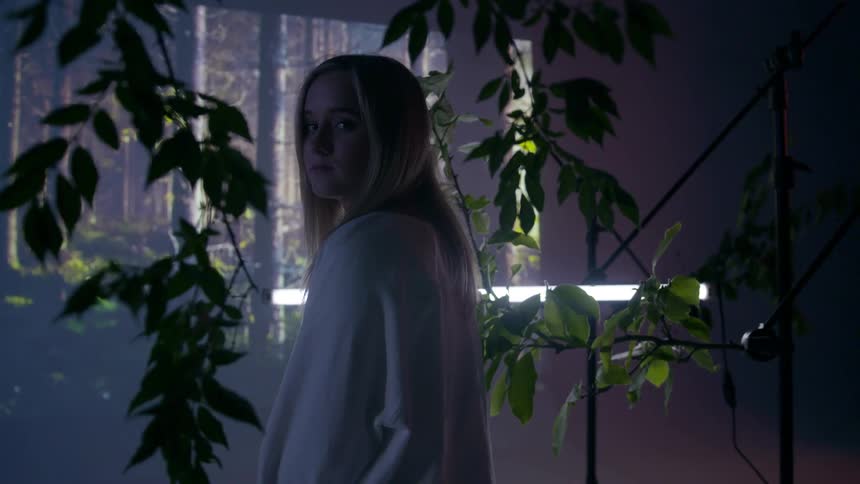 A young girl stares from inside a dim room with vines
