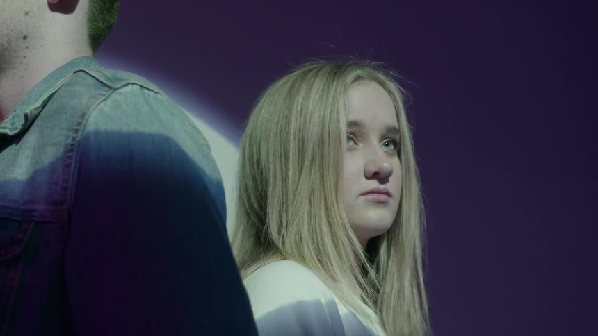 A young woman sits back to back with a young man in a room with a light
