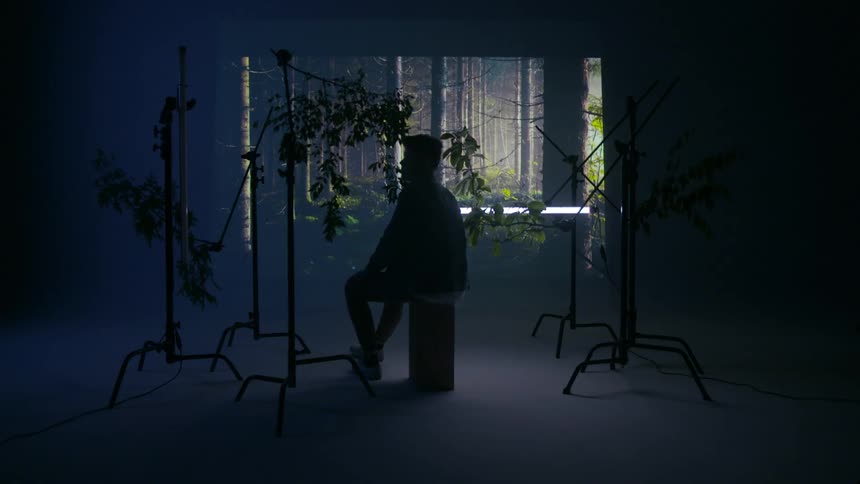A man surrounded by stands holding branches sits on a box in a studio