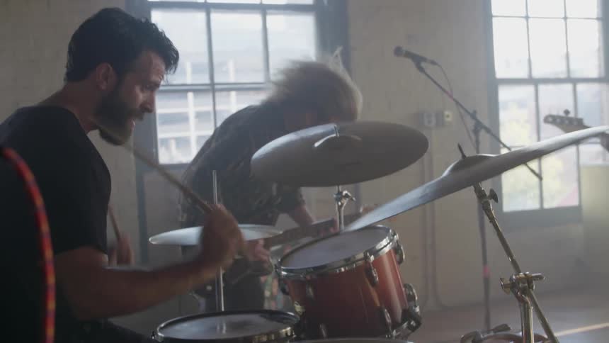 A man plays the drums while another plays a guitar