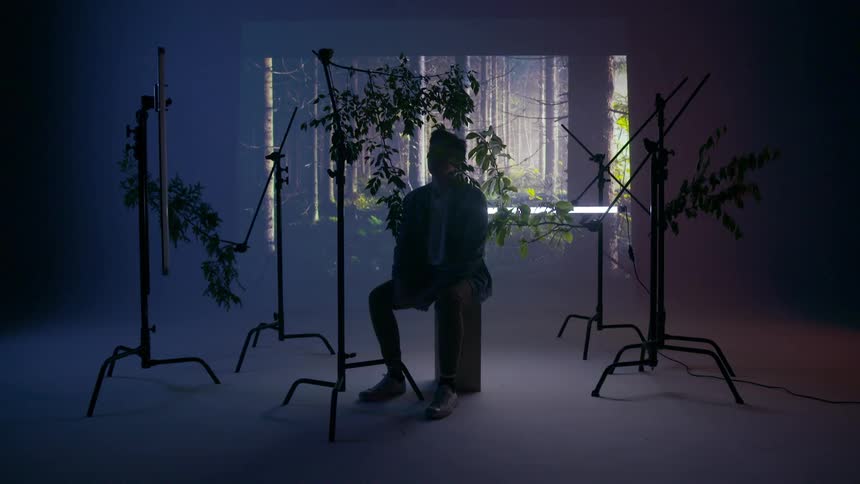 A young man sits on a box in a studio with an image projected behind him