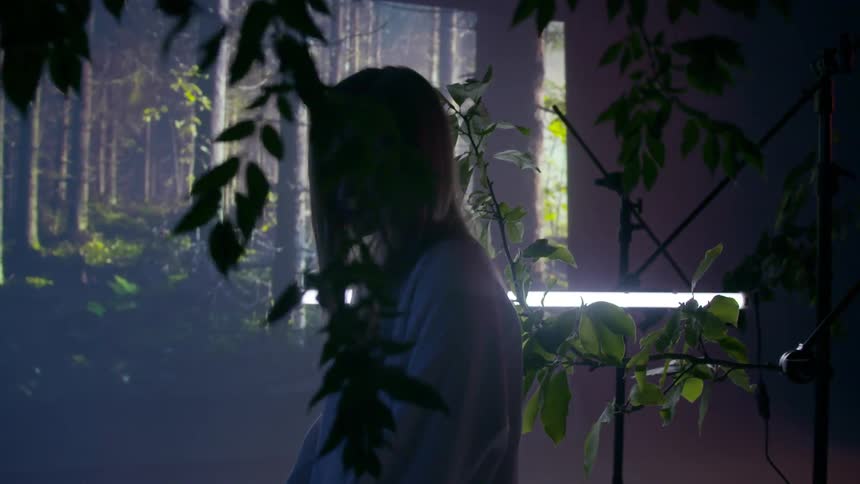 A woman is sitting unmoving in a room with tree branches and lights