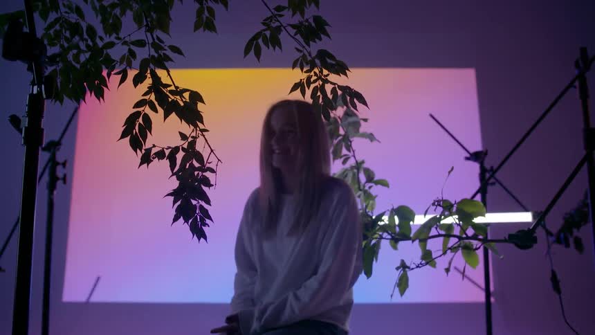 A young woman smiles as she sits in a studio surrounded by branches hand from stands