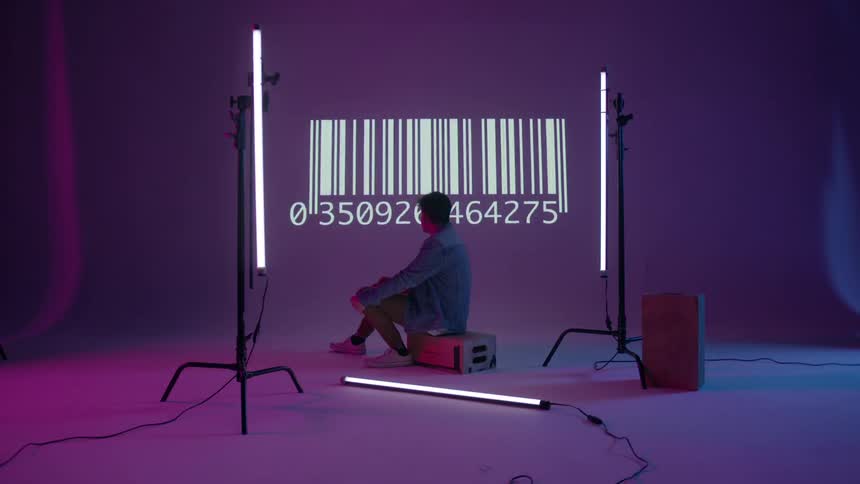 A barcode flashes on the wall behind a young man sitting on a box