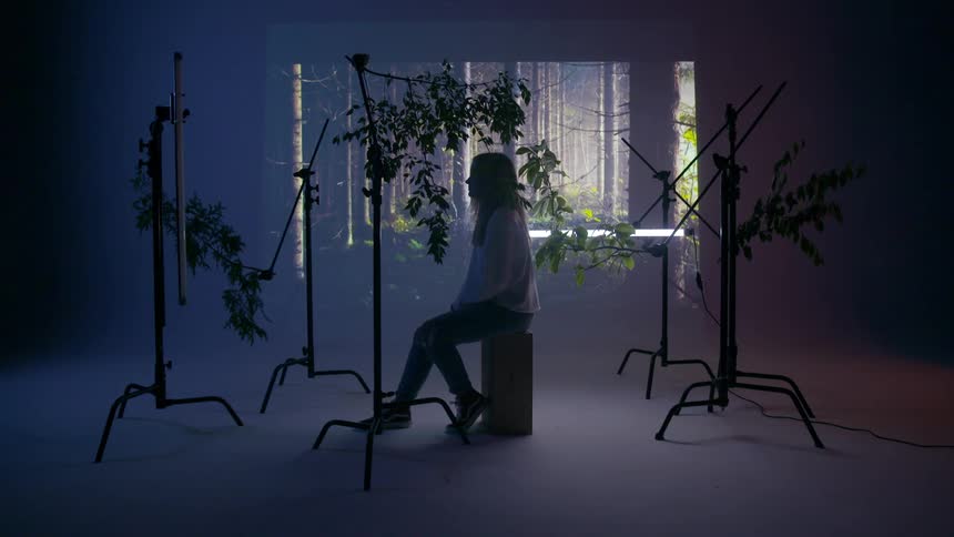 A young woman sits on a box in a studio surrounded by tree branches