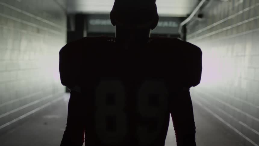 Man in a football uniform holds a helmet as we looks down a corridor