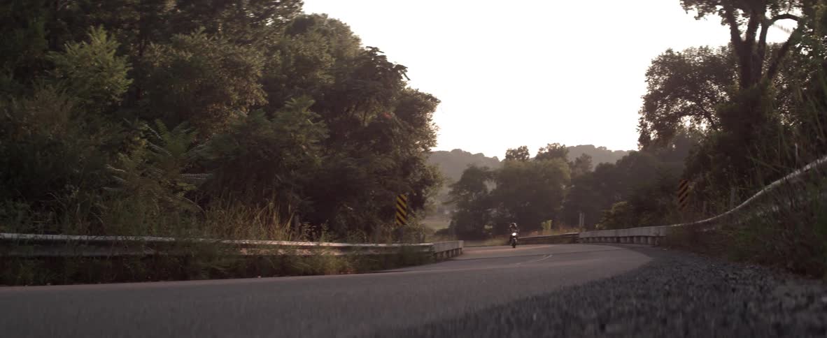 A woman is riding a motorcycle on a rural road at dusk or dawn