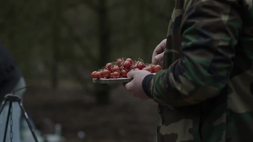 A man is holding a plate of tomatoes as he stands near a camp stove