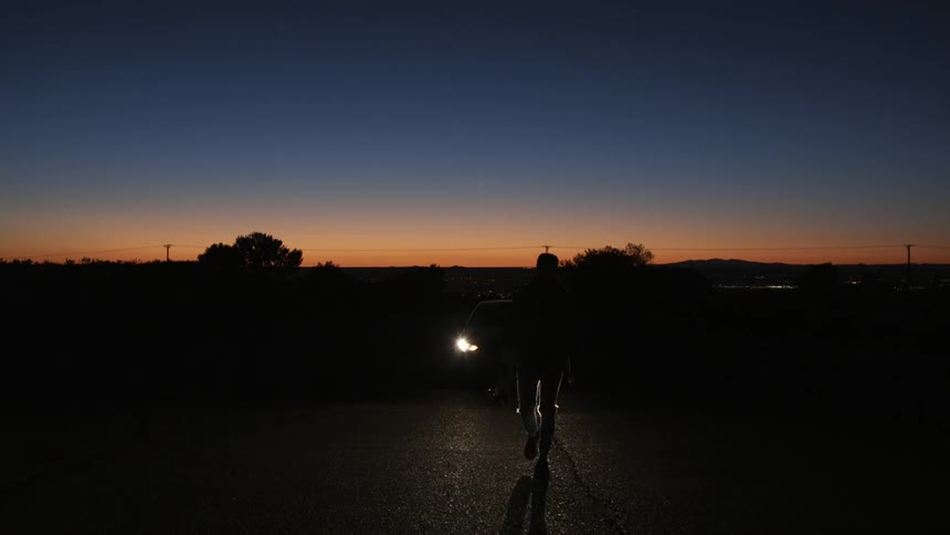 Skateboarder rides in front of car headlights at dusk