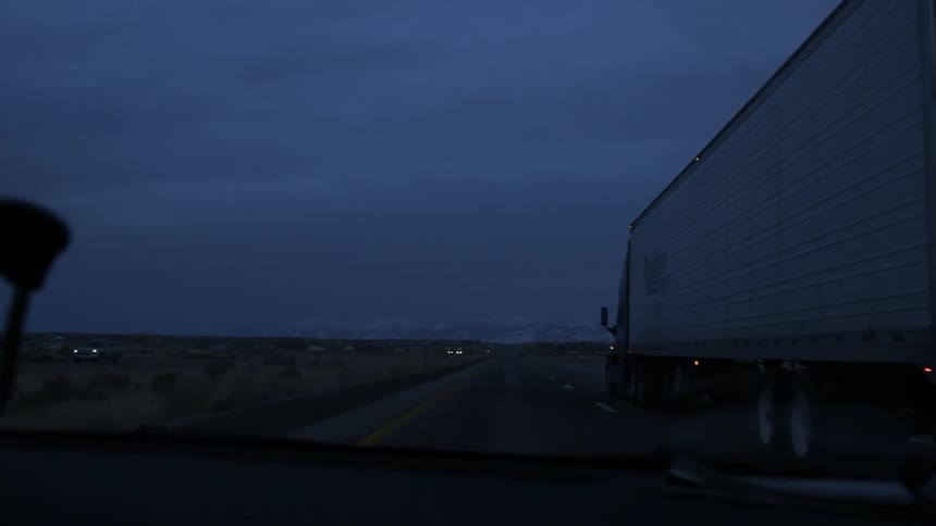 A vehicle is passing a semi-truck on a desert highway at dusk