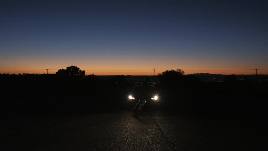 A man skateboards toward a parked car at sunset