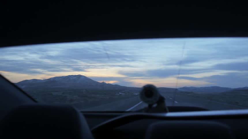 A high desert road recedes in the rearview of a car at dusk