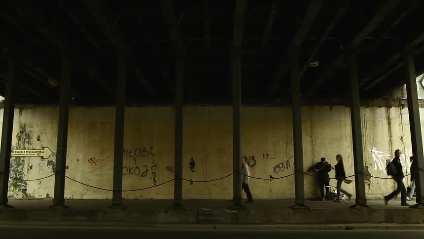 Pedestrians walk beneath a bridge as a man plays violin