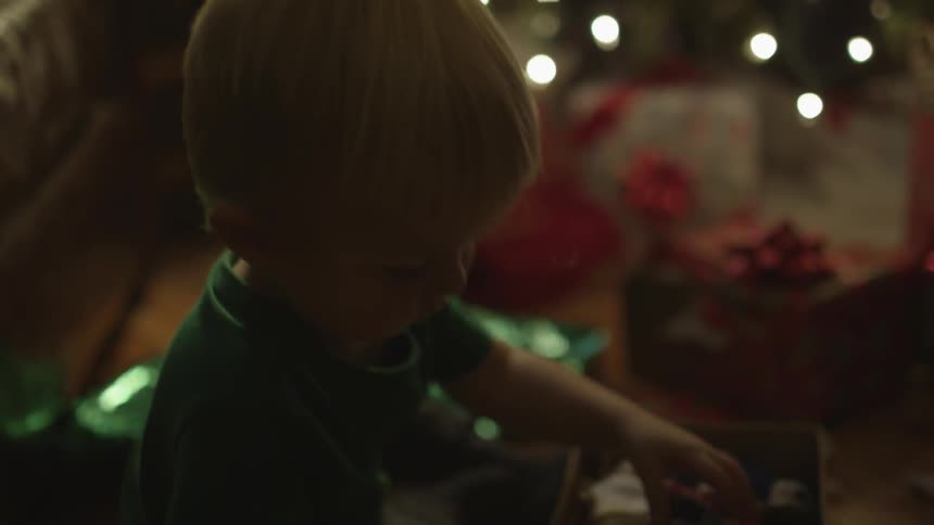 A toddler opens a gift box while seated next to a christmas tree