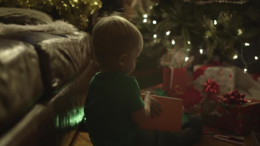 A toddler sits on the floor next to a christmas trees and gifts while flipping through a book