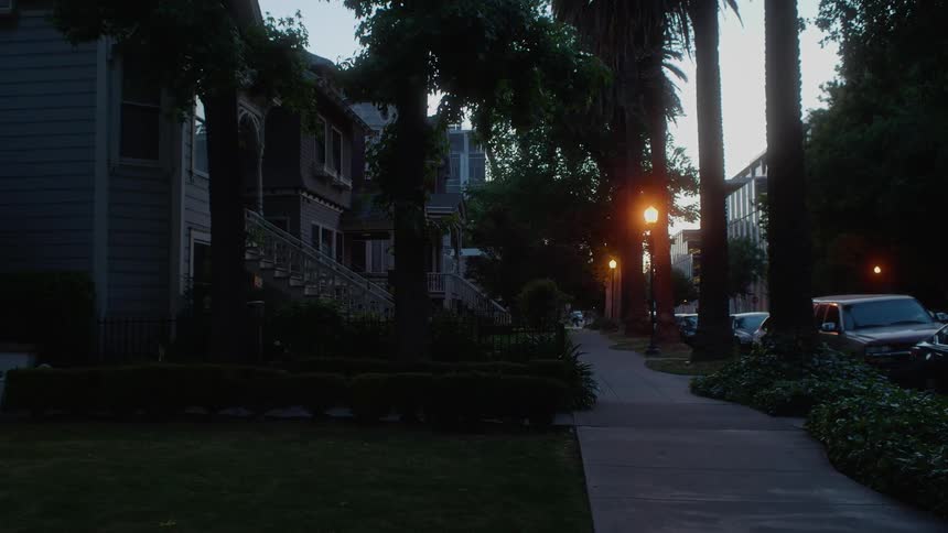 Street lamps shine along the sidewalk in a neighborhood at dawn