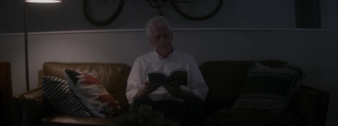 An old man sits on a couch reading a book in the evening