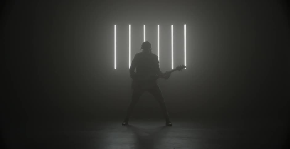 A man plays guitar in front of a wall with fluorescent tube lights