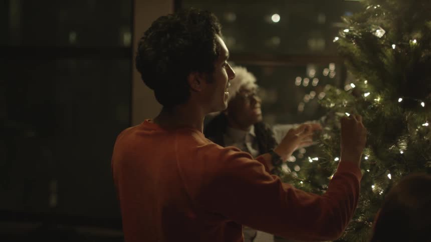 White man with a orange sweater and black woman wearing a christmas hat decorate a christmas tree at night