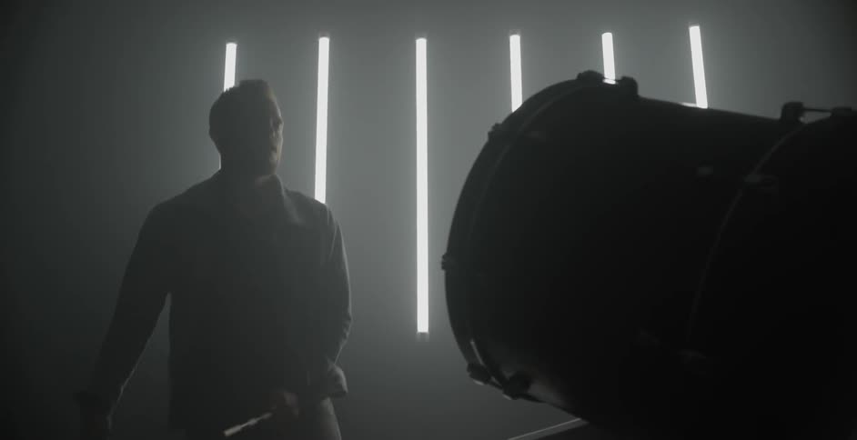 A man plays the drums in front of a wall with fluorescent light tubes