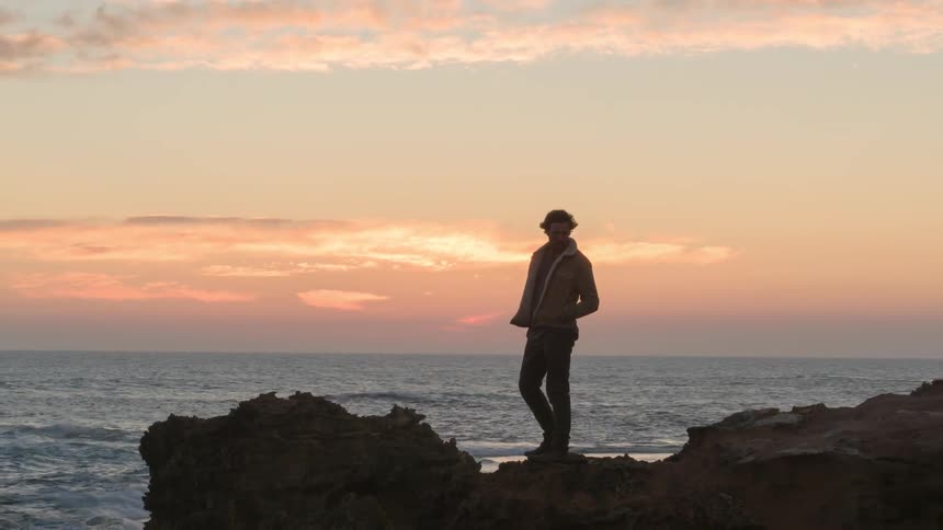 A man is standing on a rocky prominence overlooking the sea at sunset
