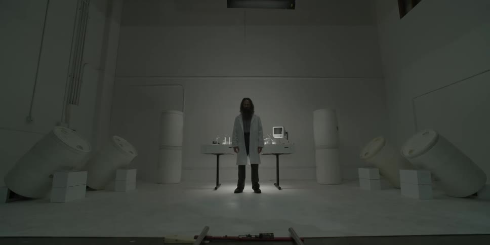 A disheveled man in a white coat stands in lab