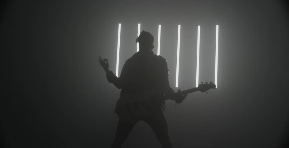 A man is playing a guitar in a dark room with a slatted window
