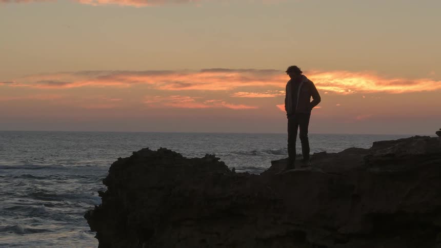 A man stands on the edge of a cliff near water