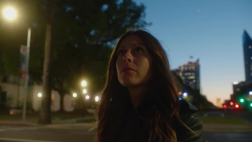 A woman standing outside in a city at dusk is looking towards the sky