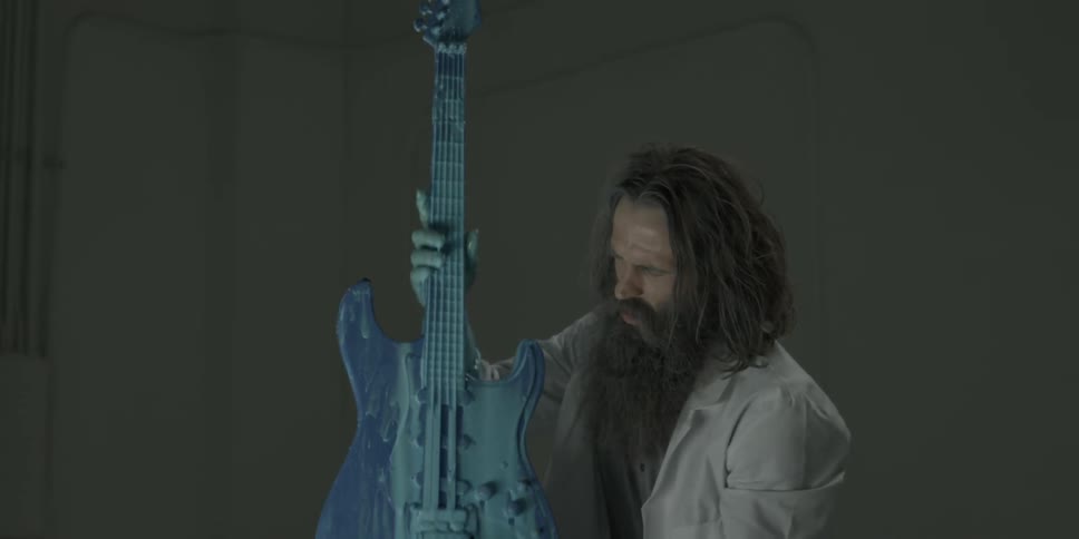 A man in a lab coat holds up a guitar covered and dripping in blue paint