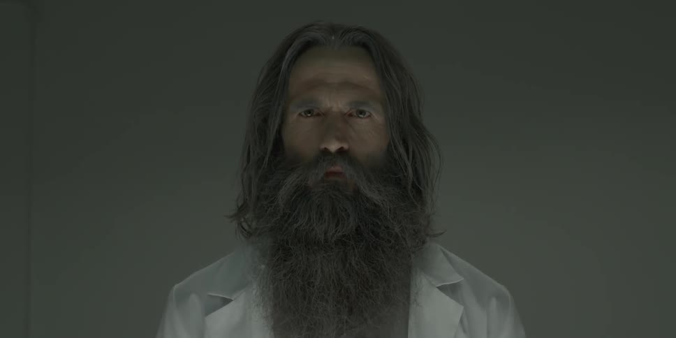 A man with long hair and beard wearing a lab coat stares straight ahead